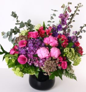 A stunning mix including peonies, hydrangea, delphinium and more is designed in a black glass vase.