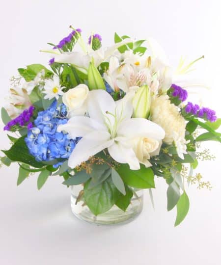 An assortment of blue and white flowers, including blue hydrangea, white roses or spray roses, and other long lasting options arranged in a glass cylinder vase. 