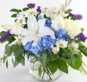 An assortment of blue and white flowers, including blue hydrangea, white roses or spray roses, and other long lasting options arranged in a glass cylinder vase.